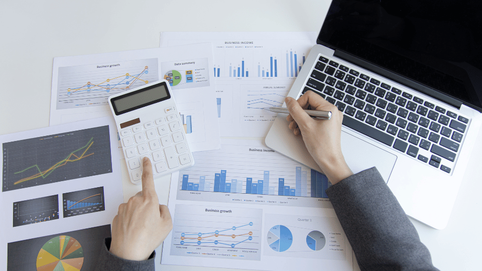 Manos utilizando una calculadora junto a gráficos y un portátil que muestran datos de crecimiento empresarial, representando el análisis de capital de trabajo.