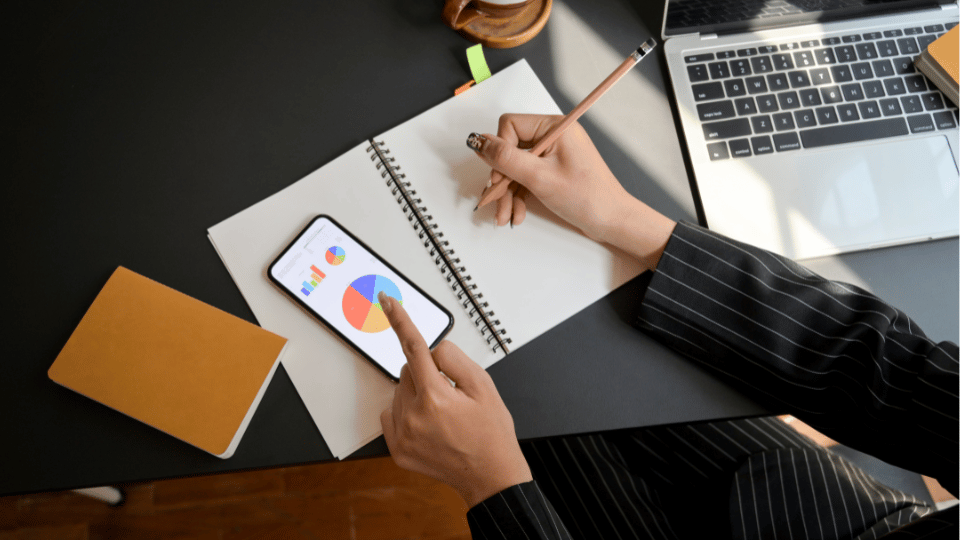 Person analyzing revenue charts on a smartphone and notebook, with a laptop in the background, representing data-driven decision-making in revenue-based financing.