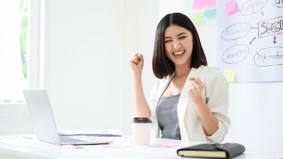 A smiling entrepreneur celebrating success at her desk with a laptop, coffee, and business strategy notes in the background, representing the positive impact of revenue-based financing.