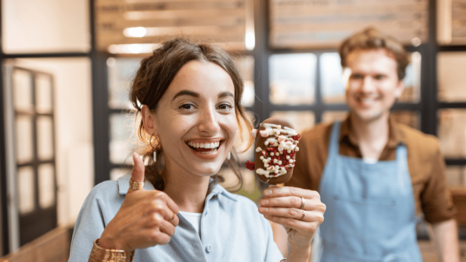 A happy customer giving a thumbs up while holding a dessert, with a smiling vendor in the background. The image represents gathering customer feedback and satisfaction, showcasing the importance of surveying customers to understand their preferences and experiences.