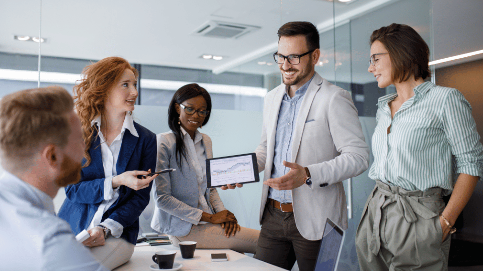 A group of five business professionals in a meeting room, discussing a sales strategy. One man holds a tablet displaying data charts, while others listen and engage in conversation.