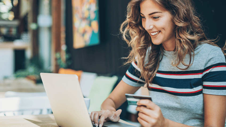 A woman sitting at a desk using her laptop and holding a credit card, representing online shopping. She is smiling, symbolizing a positive customer experience and the importance of understanding customer behavior and preferences.